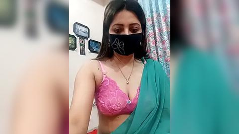 Media: Video of a young woman with medium skin tone, wearing a black face mask, a pink lace bra, and a green sari. Background includes a wall with framed photos and a colorful curtain.