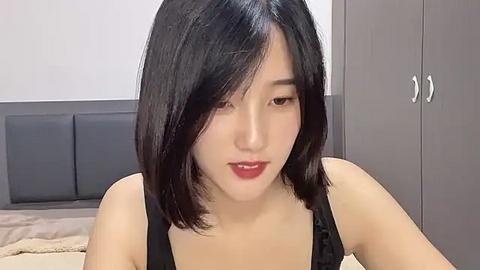 Media: Video of a young East Asian woman with straight black hair, fair skin, and light makeup, wearing a black top. She sits on a bed with a beige headboard and gray wardrobe in the background.