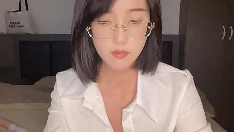 Media: Video of an Asian woman with shoulder-length black hair, wearing round glasses, a white blouse, and earphones, seated in a dimly lit bedroom with a bed and dark furniture in the background.