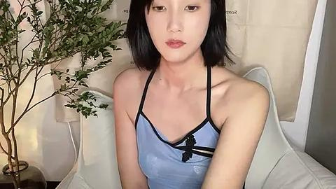 Media: Video of an East Asian woman with straight black hair and light skin, wearing a blue spaghetti strap top with black trim, sitting on a white couch with a beige wall and potted plant in the background.