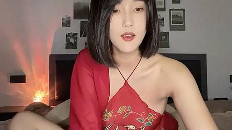 Media: Video of an Asian woman with straight black hair, wearing a red, floral-patterned off-shoulder top, sitting on a bed with a dark headboard and beige bedding. The background features a dimly lit room with a candle and framed photos.