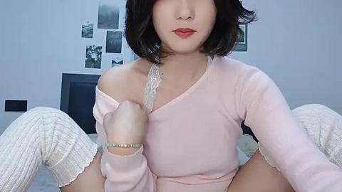 Media: Video of an East Asian woman with fair skin, wearing a pink off-shoulder sweater, black lace bra, and white ribbed socks, posing in a bedroom with a white wall adorned with framed photos.