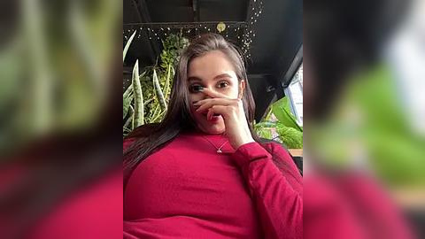 Media: Video of a plus-sized woman with long dark hair, wearing a red sweater, partially covering her face with her hand, in a green, indoor setting with potted plants.