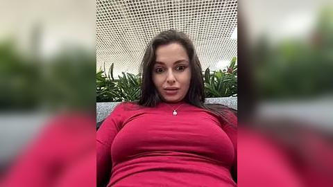 Media: Video of a woman with long dark hair, wearing a red long-sleeved top, sitting in a modern, light-colored room with a grid ceiling and green plants in the background.