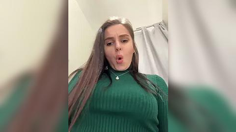 Media: A video of a young woman with long, straight brown hair, wearing a green sweater, sticking her tongue out, in a bathroom with a white shower curtain.