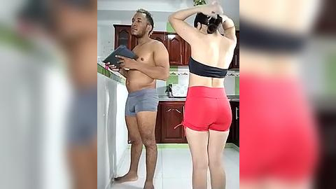 Media: Video of a muscular, shirtless man wearing gray shorts, standing beside a woman in a black sports bra and red shorts, in a modern kitchen with wooden cabinets and white countertops.