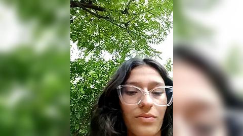 Media: Video of a woman with long black hair and clear glasses, standing in a lush, green forest, surrounded by vibrant foliage. Her expression is neutral, and she appears to be wearing a black top.