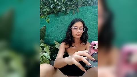 Media: Video of a young woman with medium skin tone, long black hair, wearing glasses and a black strapless top, seated outdoors, holding a pink phone, surrounded by greenery and foliage.