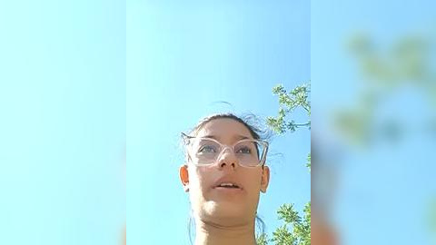 Media: Video of a light-skinned, young woman with glasses, looking upward, against a clear blue sky with green foliage in the background.