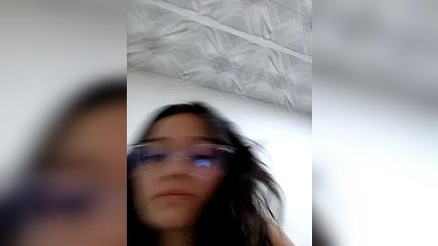 Media: Video of a young woman with long, dark hair wearing clear glasses, standing in a room with a textured, white ceiling. The background is slightly blurred.