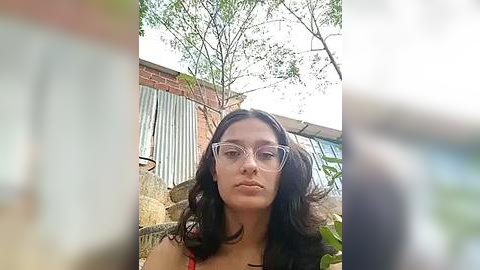 Media: A video of a young woman with long, dark hair, wearing clear-framed glasses and a red dress, standing in a suburban backyard with trees, a brick wall, and a metal gate in the background.