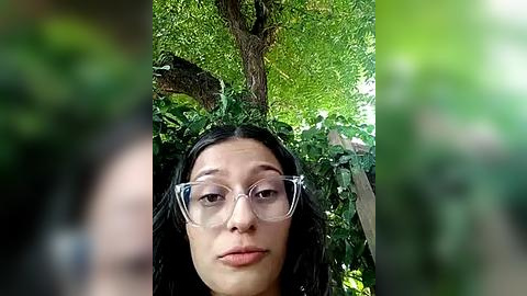 Media: Video of a young woman with long black hair, wearing clear glasses and a black top, standing in front of a lush green forest with a large tree in the background.