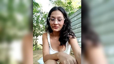 Media: Video of a young woman with long black hair and glasses, wearing a white tank top, sitting outdoors with blurred greenery and a corrugated metal structure in the background.