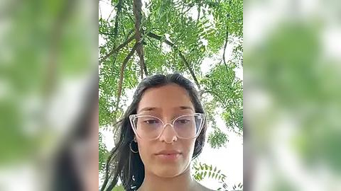 Media: Video of a young woman with long, dark hair and clear glasses, standing in a lush, green forest. The background is blurred, with sunlight filtering through dense foliage.