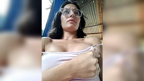 Media: Video of a woman with glasses and dark hair, wearing a white tank top, partially covering her breasts with her hand. Background shows a blue wall and metal structure, giving an industrial vibe.