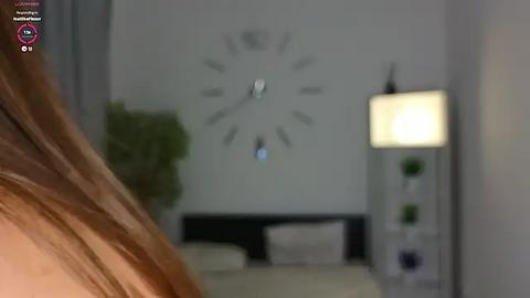 Media: Blurry photo of a modern, minimalist bedroom with a large wall clock, a lamp, and a bed. The background features a green plant.