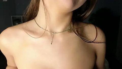 Media: Video of a topless woman with light skin and long brown hair, wearing a delicate gold chain necklace. The background is dark and indistinct.