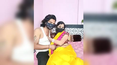 Media: Video of two women in a hospital room; one in a white tank top, black mask, and green pants, the other in a yellow sari, black mask, and a black top, seated on a pink bed.