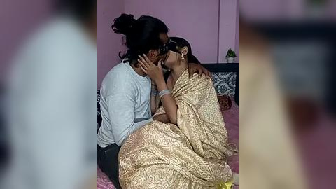 Media: A video shows a woman in a gold sari kissing a man in a light gray shirt on a pink bed. The background features a pink wall and a potted plant.