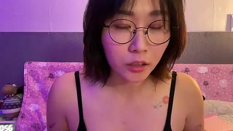 Media: Video of an Asian woman with short, dark hair and glasses, wearing a black spaghetti strap top, lying on a bed with pink floral sheets, against a purple-lit wall.