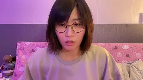 Media: Video of an East Asian woman with shoulder-length brown hair, wearing round glasses, a light-colored sweater, and a slightly pensive expression. She sits on a bed with a pink floral-patterned blanket and stuffed toys in the background.