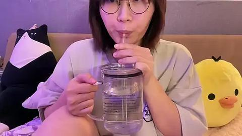 Media: Video of an Asian woman with glasses and straight black hair, wearing a light gray shirt, drinking from a glass jar, surrounded by plush toys.