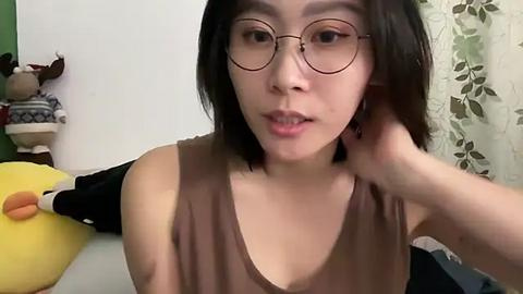 Media: Video of a young Asian woman with straight black hair, wearing glasses, and a brown sleeveless top, sitting on a bed. Background includes a green wall, a stuffed animal, and a floral curtain.