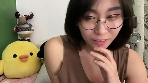 Media: Video of an East Asian woman with glasses, short black hair, and a brown sleeveless top, touching her chin, in a cozy room with a plush yellow duck toy, a green wall, and patterned curtains.