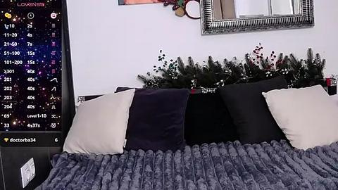 Media: A video of a cozy bedroom with a bed covered in a plush, gray blanket, adorned with a black and white pillow set. A starry night sky digital clock and festive greenery decorate the white wall background.