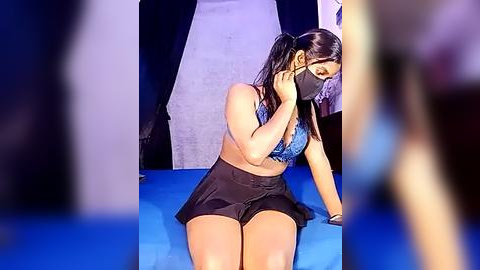 Media: Video of a woman with medium skin tone, wearing a blue lace bra and black shorts, kneeling on a blue carpet, in a fashion runway setting.