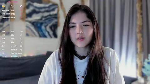 Media: Video of a young Asian woman with long black hair, wearing a white robe, standing indoors in a dimly lit room with modern art on the wall.