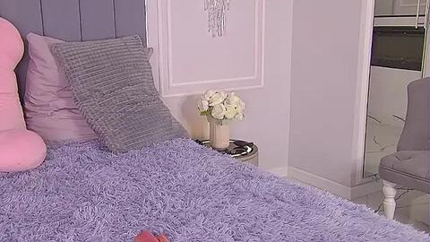 Media: A video of a pastel-themed bedroom featuring a plush lavender bedspread, gray textured pillows, and a vase of white roses on a side table against a white wall with a decorative chandelier.