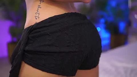 Media: Video of a woman's bare back and shoulder, showing a black shirt draped off the shoulder, revealing a tattoo with cursive text. Background is blurred, featuring blue and purple lighting and green plants.