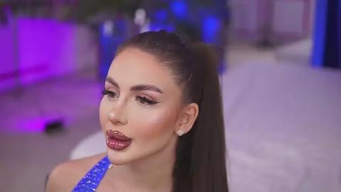 Media: Video of a light-skinned woman with long, dark hair pulled back, wearing heavy makeup with bold eyebrows, pink eyeshadow, and red lipstick. She's wearing a blue sequined top, with a blurred, purple-lit indoor background.