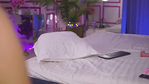 Media: Video of a plush white pillow on a neatly made bed, with a remote control and a pink object nearby, in a dimly lit room with a purple accent, featuring a large potted plant and a partially visible person.