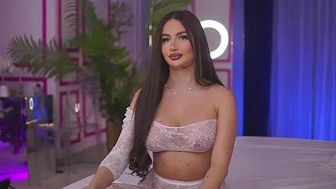 Media: Video of a fair-skinned, long-haired woman in a white, lace bralette and matching panties, seated on a bed in a pink, modern room with a round mirror and green plants.