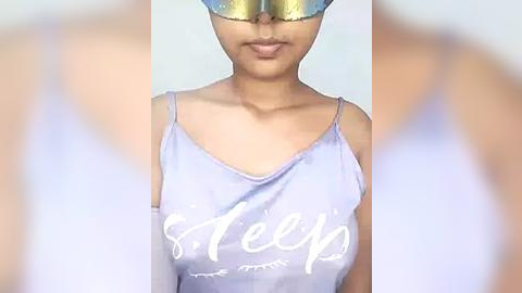 Media: Video of a woman with medium skin tone wearing a light purple spaghetti-strap tank top with \"Sleep\" written in white cursive and a golden sequin eye mask. The background is blurred, focusing on the subject's upper body.