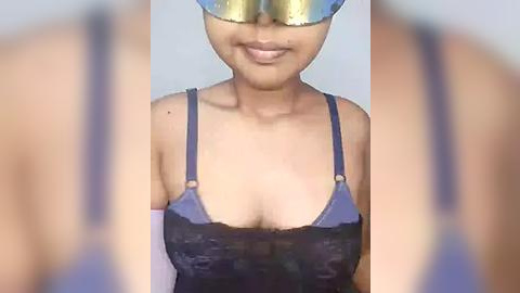 Media: Video of a woman with medium brown skin and a slender physique wearing a dark blue lace bra, gold face mask, and white elbow-length gloves, against a blurred background.