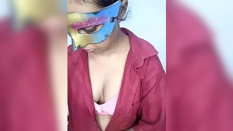 Media: Video of a young Asian woman with medium skin tone wearing a pink bra and red unbuttoned shirt, her face partially obscured by a colorful, reflective mask. Background is blurred, focusing attention on her exposed cleavage.