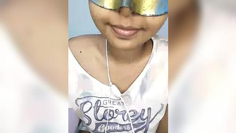 Media: Video of a young Asian woman with light skin, wearing a white tank top with \"Great Story Goddess\" text, gold reflective sunglasses, and a white necklace. The background is blurred.