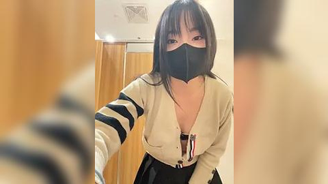 Media: A video of an Asian woman with straight black hair and fair skin wearing a beige cardigan, black mask, and striped sleeves, standing in a room with wooden walls and a ceiling vent.