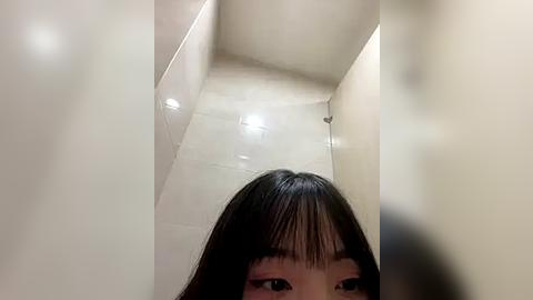 Media: Video of a young Asian woman with straight black hair and bangs, partially visible in a narrow, dimly lit, beige-tiled bathroom, with a blurred reflection of a person in the foreground.