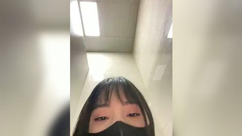 Media: A video of a young Asian woman with straight black hair and bangs, wearing a black mask, taken from below. She stands in a brightly lit, white-walled hallway with fluorescent lights and clean, tiled floors.