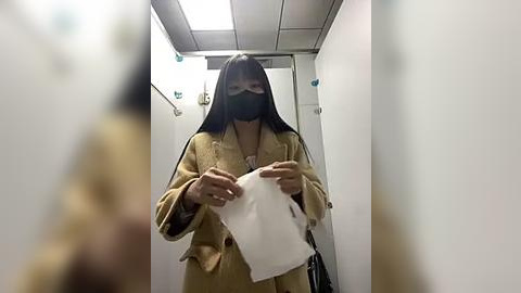 Media: A video of a person in a beige coat and black face mask holding a white tissue in a white-tiled hallway with fluorescent lighting.