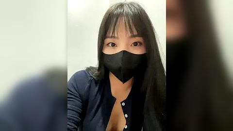 Media: Video of an East Asian woman with straight black hair and medium-length bangs, wearing a black face mask, dark blue button-up shirt, and a black cardigan. She has a slight smile, and the background is blurred.