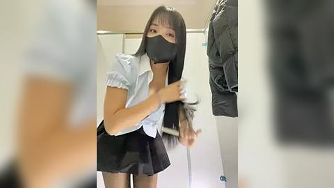 Media: Video of an East Asian woman with long black hair, wearing a white blouse, black skirt, black face mask, and black gloves, standing in a dressing room, adjusting her hair.