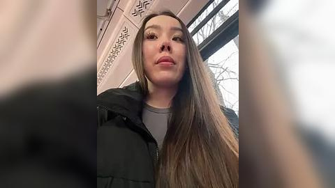 Media: Video of a young Asian woman with long, straight brown hair, wearing a black jacket, standing in a subway car. The background features subway window and ornate white wall patterns.