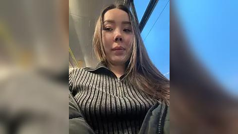 Media: A video of a young Asian woman with long, straight, light brown hair, wearing a striped, sleeveless top and a black jacket, seated on a bus with a blue sky and blurred buildings in the background.