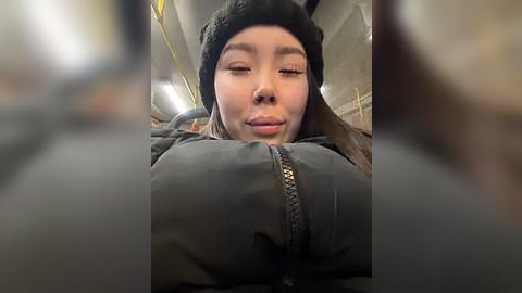 Media: A video of an East Asian woman with straight black hair, wearing a black beanie and a dark jacket, resting her head on her hand, looking contemplative in a subway car with yellow seats and overhead lights.