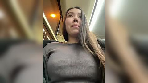 Media: Video of an Asian woman with long, straight black hair, wearing a tight, grey ribbed sweater that accentuates her large breasts, sitting in a car.